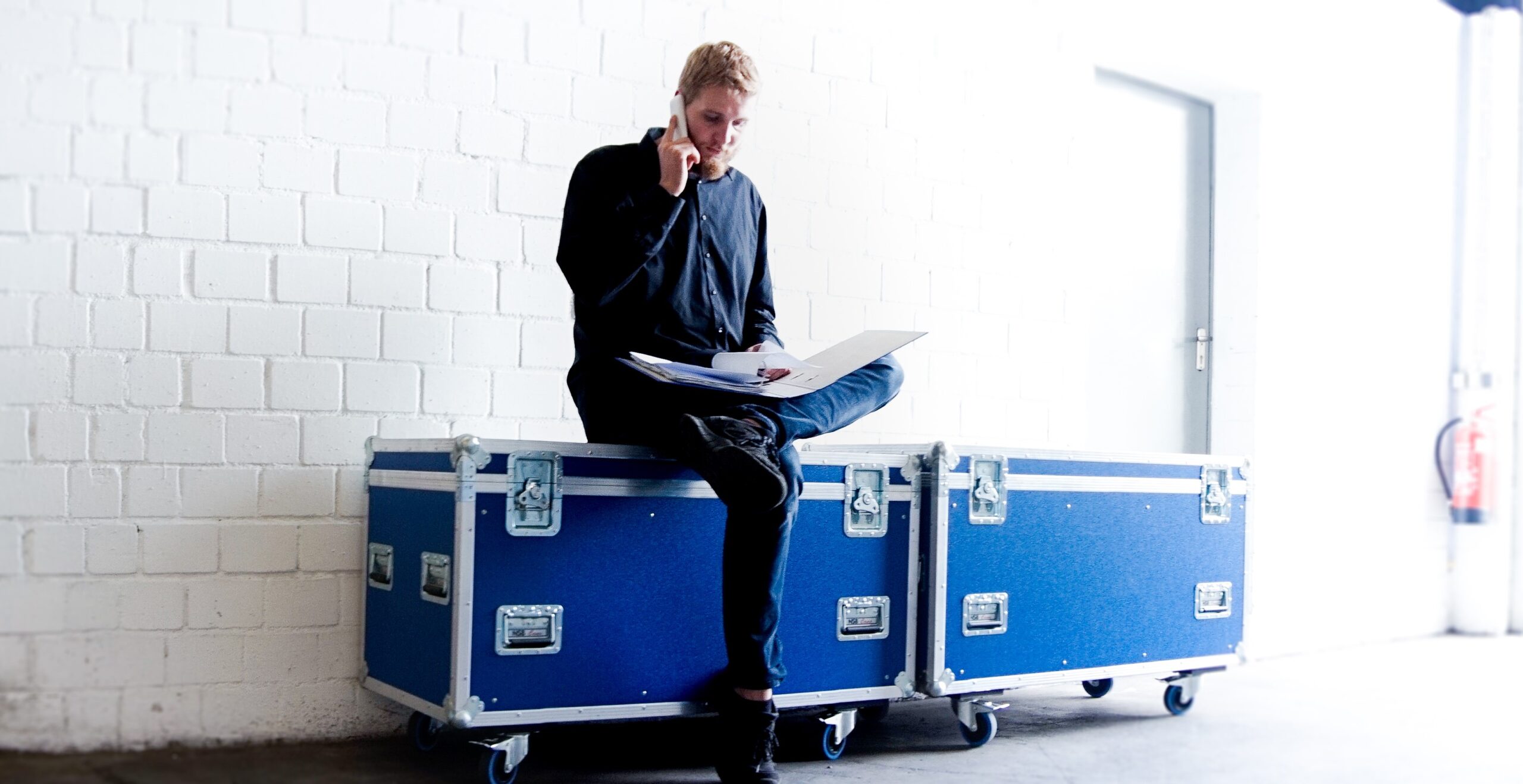 Projektmanager und Geschäftsführer Felix Nüske sitzend auf einem blauen Case, Telefoniert und schaut in einen Aktenordner.