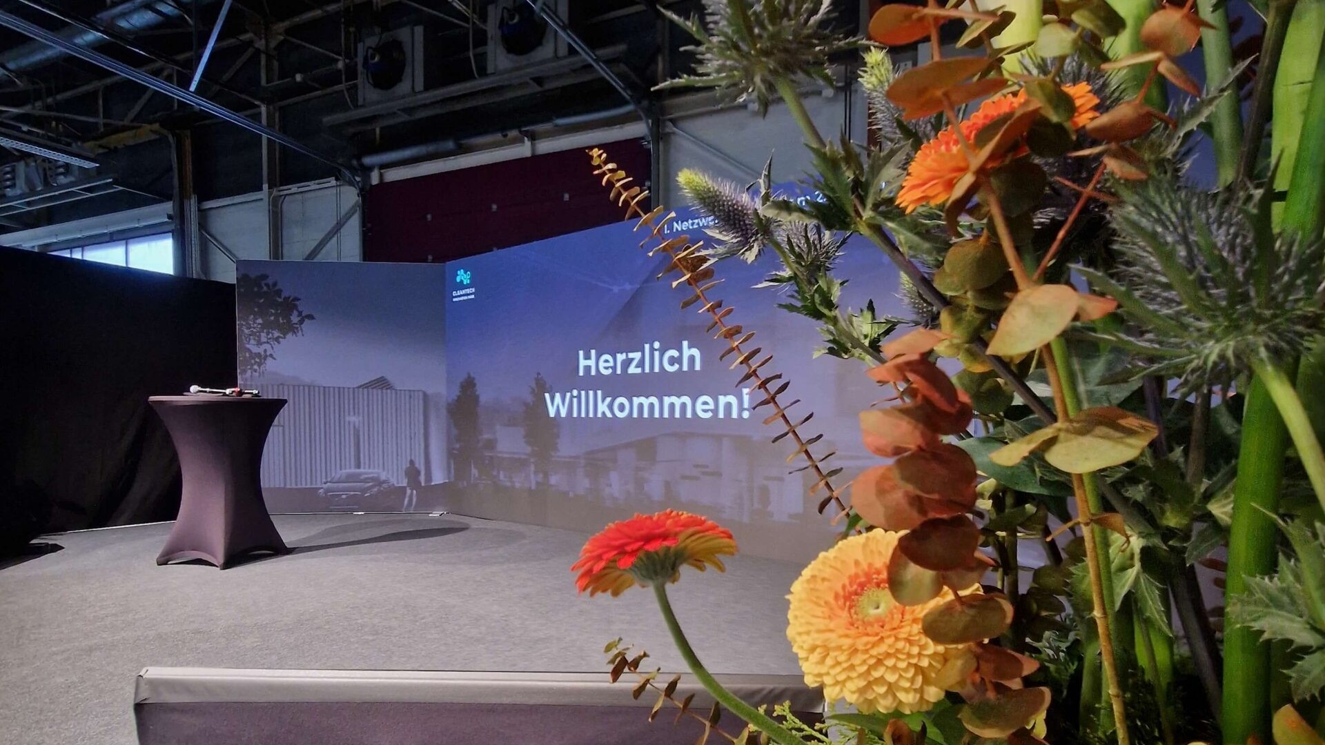 LED-Wand auf Bühne. Zu sehen ist eine Präsentation. Der Text sagt: Herzlich Willkommen. Im Vordergrund ist ein Blumenstraus.